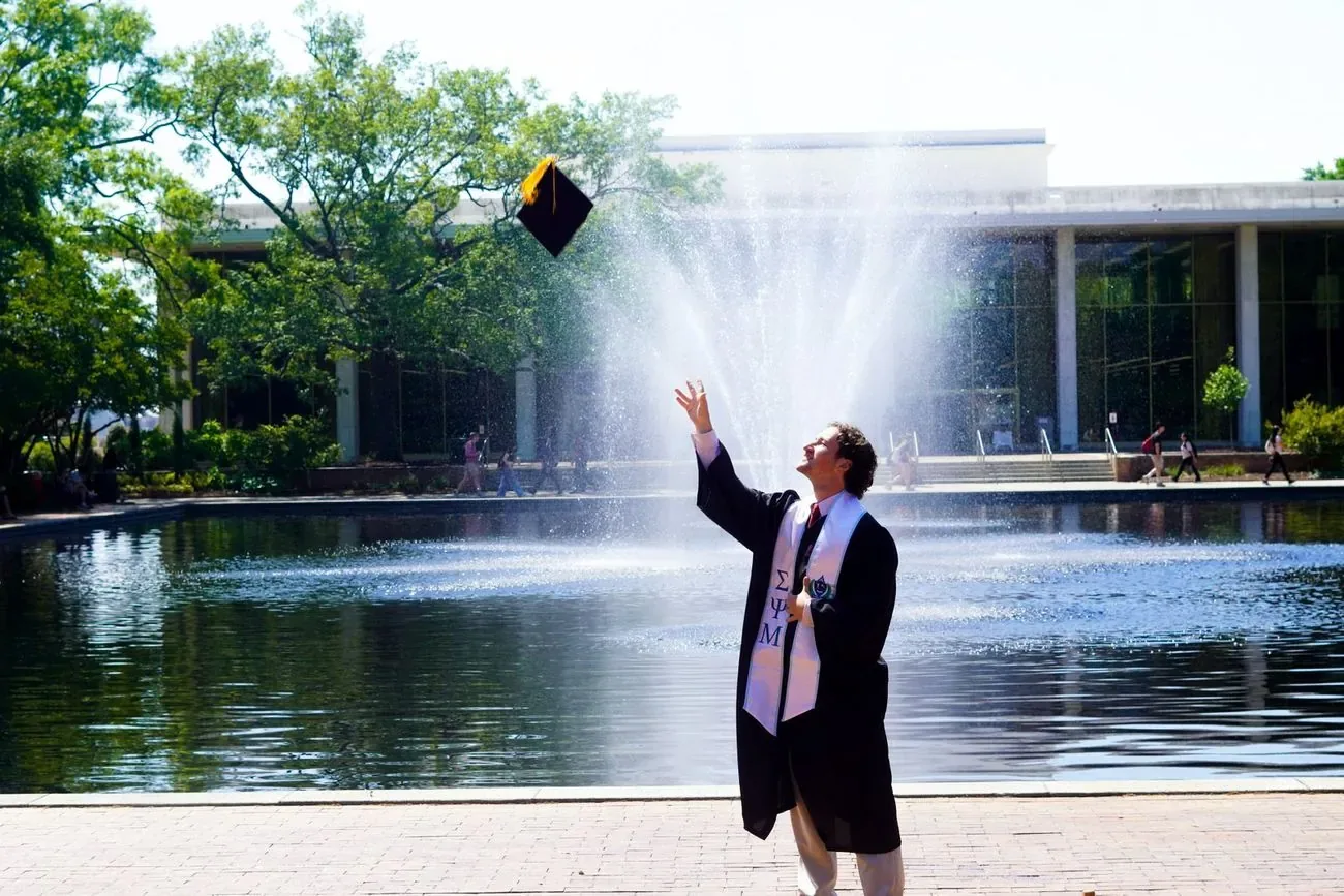Graduate in cap and gown tossing a mortarboard in the air beside a campus fountain