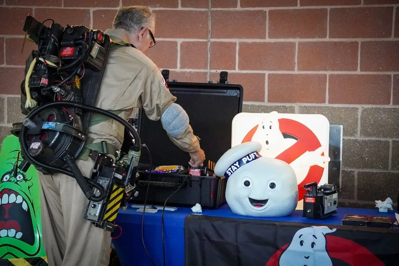 A costumed Ghostbuster with a proton pack setting up a display table on theme night