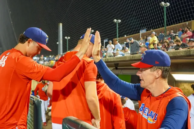 Coach in an orange jacket high-fiving players over the dugout rail