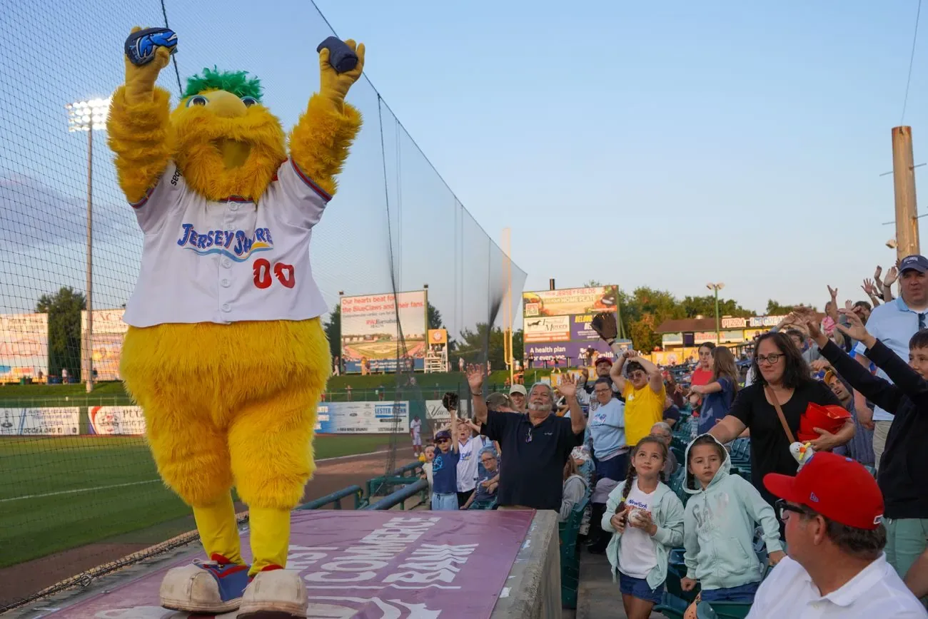Buster the mascot standing on the dugout roof with both arms raised over cheering fans