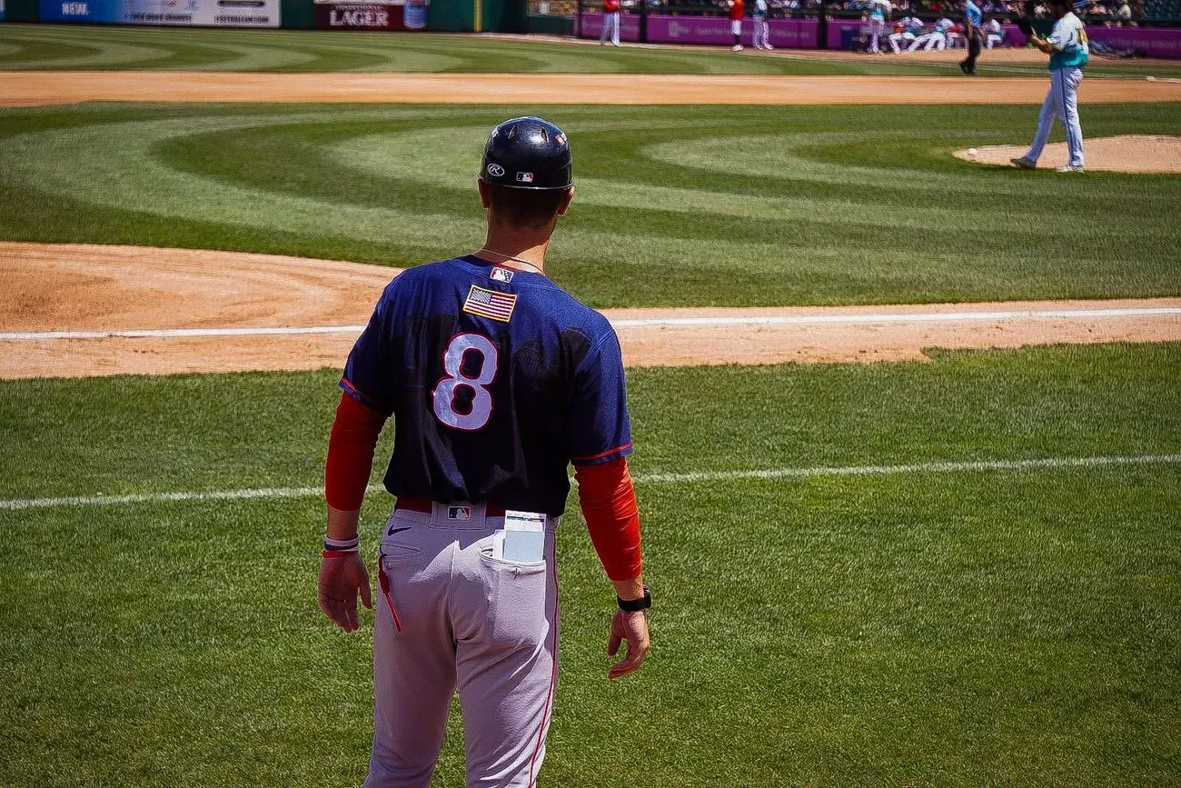 Coach in a navy jersey watching the pitcher from down the first base line