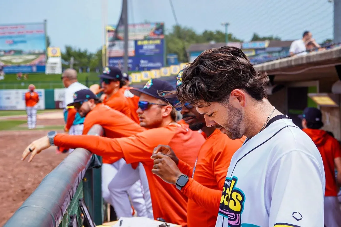 Player in a teal jersey signing autographs along the dugout rail as teammates lean over