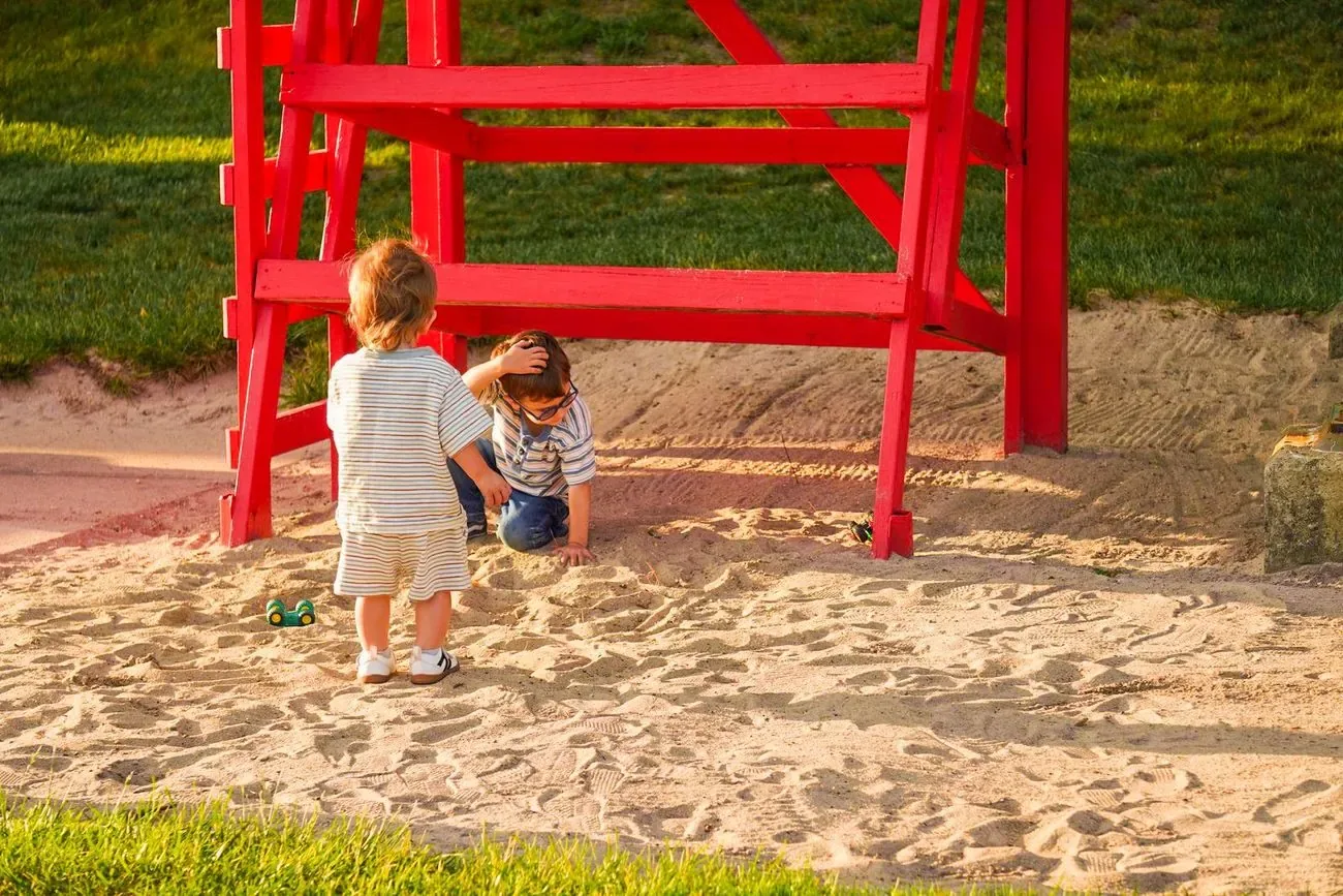 Two small kids playing in the sand under a red lifeguard chair