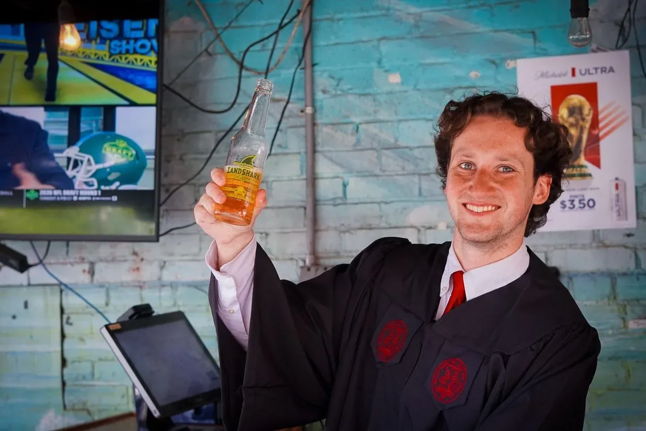 Graduate still in his gown raising a drink at a bar
