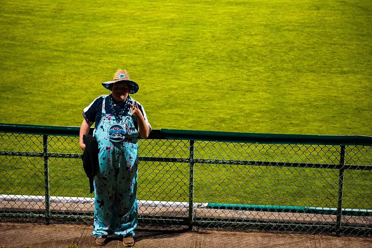 Fan in a wide-brim hat and patterned dress leaning on the outfield fence against deep green grass