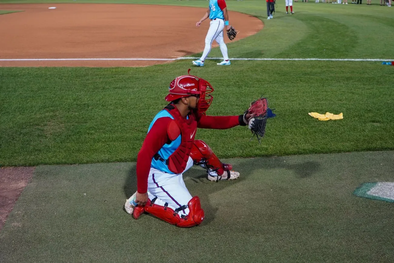 Catcher in red gear crouched in the outfield grass warming up a pitcher
