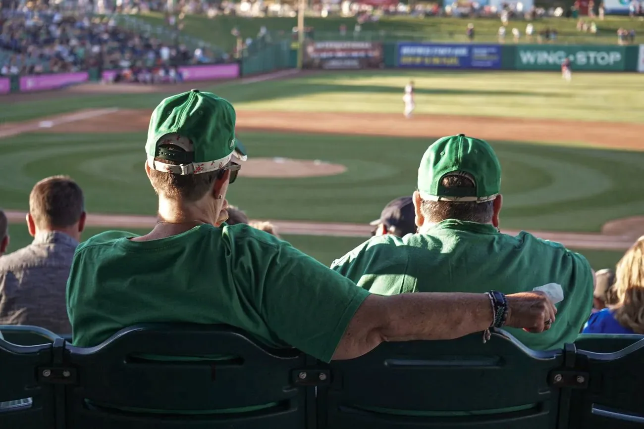 Two fans in matching green caps, arms around each other, watching the game from the seats