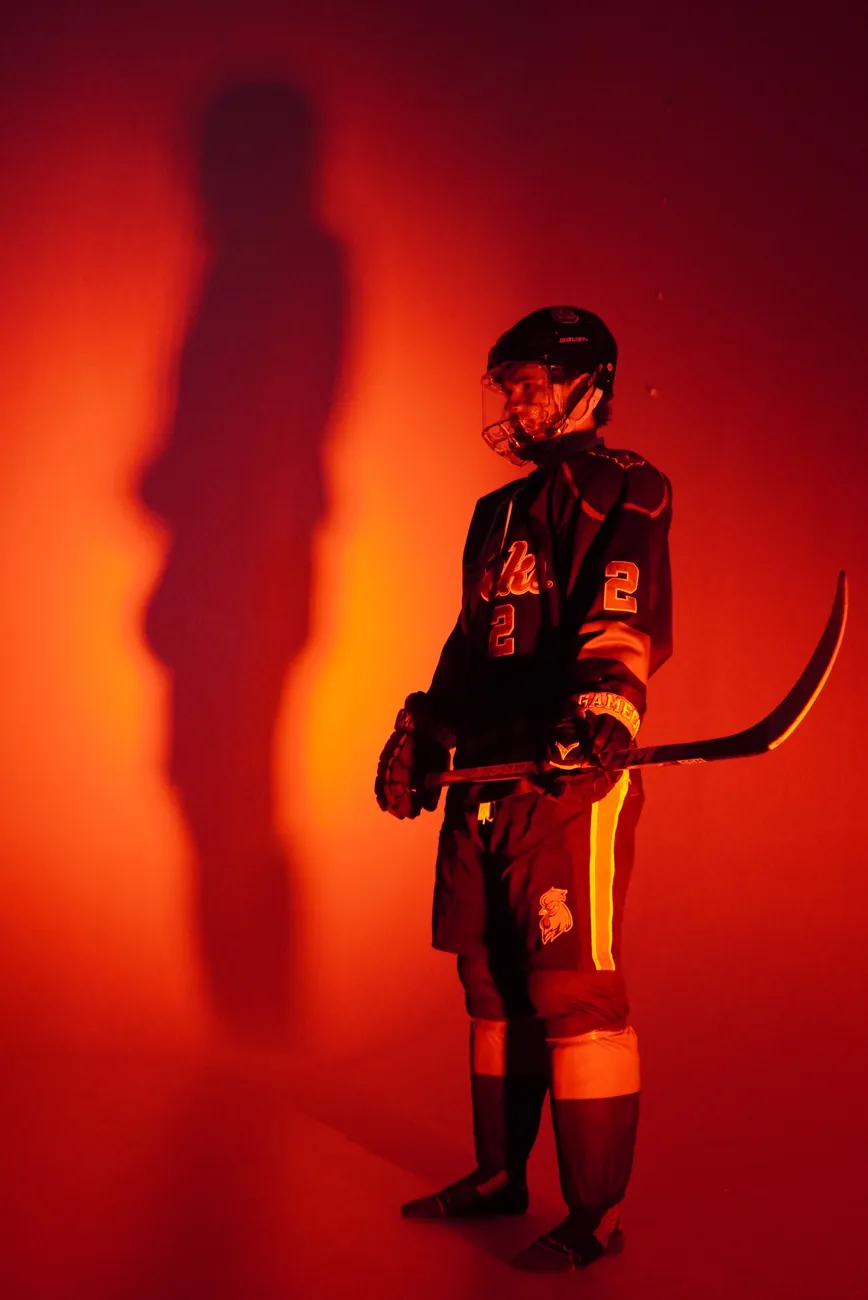 Full-length portrait of Gamecocks player number 2 holding his stick, his shadow thrown long across the red wall