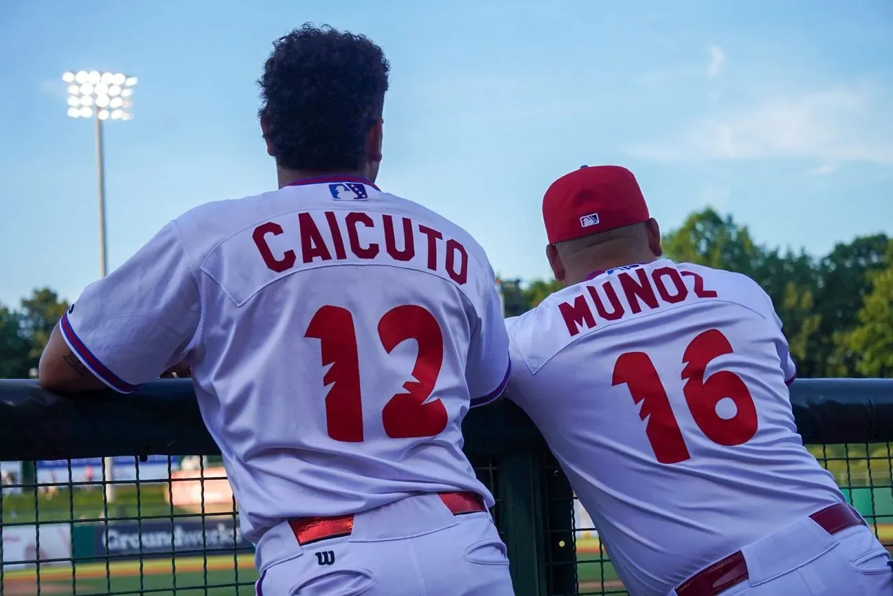 Players Caicuto and Munoz leaning on the dugout rail watching the game under the lights
