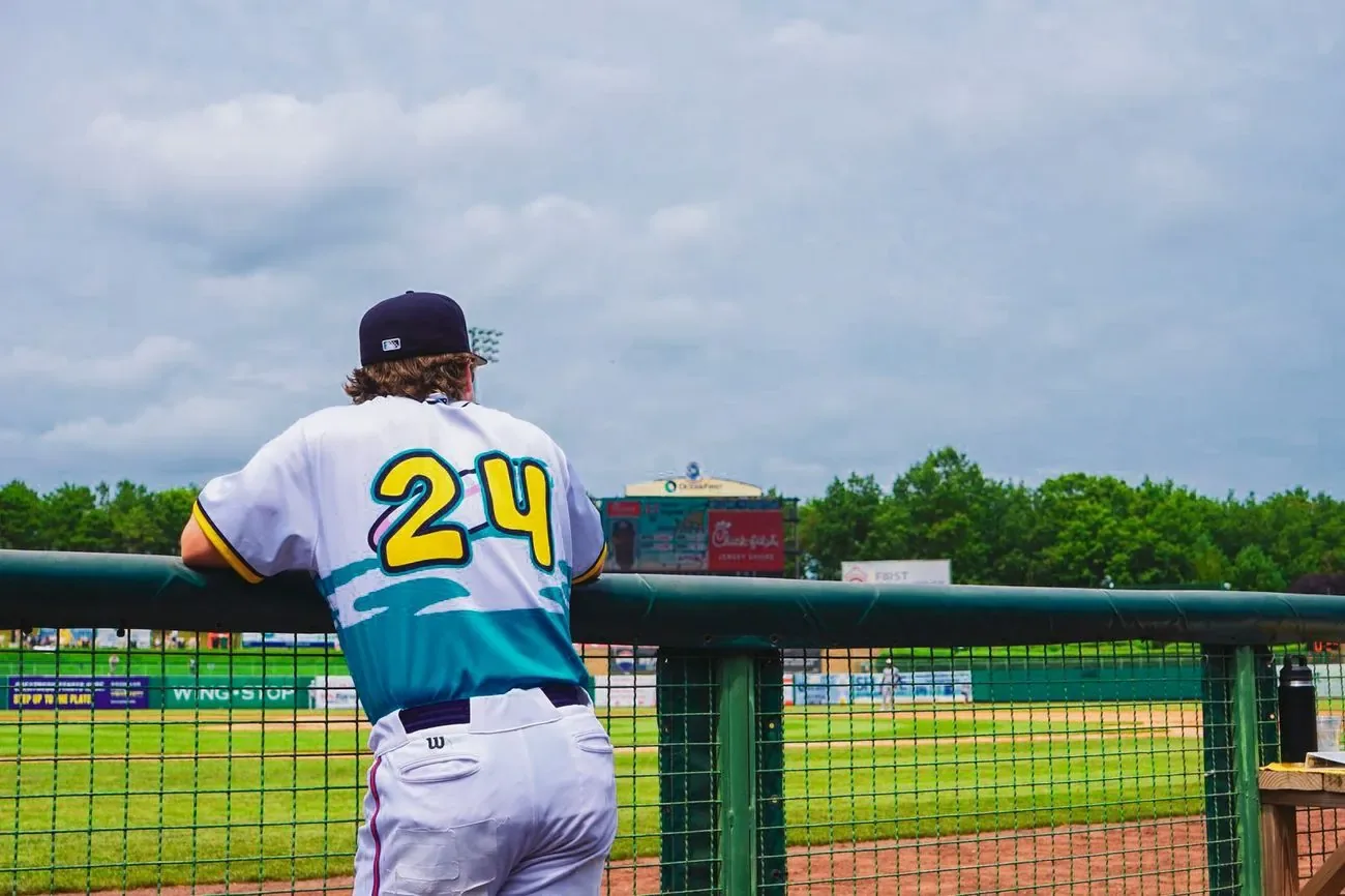 Player in a teal and yellow alternate jersey, number 24, leaning on the outfield rail in afternoon light