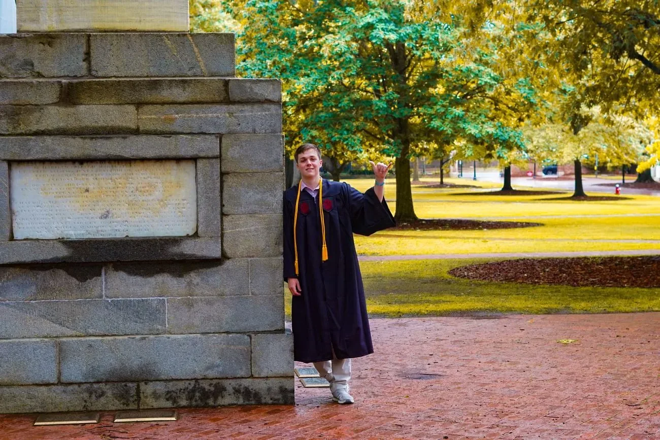 Graduate in cap and gown throwing a shaka beside a stone campus wall